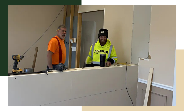 A-Verdi Facility Renovation Two A-Verdi team members working indoors on a construction project, standing behind a partially built drywall structure. One wears an orange shirt with suspenders and a headlamp, while the other wears a bright yellow A-Verdi hoodie and a winter hat, smiling with a coffee tumbler nearby. Power tools and building materials are visible around them.